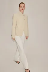 Blazer de corte estructurado en color beige, con cuello mao y cierre frontal cruzado mediante botones. Presenta mangas largas con detalle de botones en los puños y bolsillos de vivo en el frente.