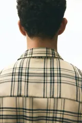 Camisa de manga larga con cuello inglés, tapeta lisa y bolsillo superior de parche. Presenta un patrón de cuadros en tonos beige claro y negro.