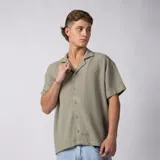 Camisa de manga corta color verde militar, confeccionada en lino entramado, con cuello cubano y cierre frontal con botones.