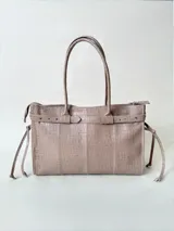 Bolso grande de cuero marrón con textura tipo cocodrilo, estructura rígida y asas para llevar al hombro. Tiene una tira con tachas y hebillas en cada extremo, también lleva lazos en los costados.