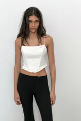 Top crop corsetero negro de popelín con escote recto, tirantes finos ajustables con lazada y bajo acabado con volante. Tiene forro interior a tono y cierre de cremallera en la espalda.
