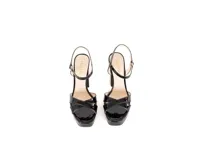 Sandalias negras de charol con plataforma y tacón ancho de 10 cm. Tiras cruzadas envolventes con hebilla dorada lateral y suela de cuero natural.