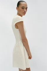 Vestido corto blanco de encaje, con cuello redondo, manga corta y bajo evasé con frunces. Cuenta con fila de falsos botones negros delanteros a contraste y cierre trasero con cremallera oculta.