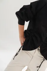 Camisa blanca de mangas largas con cuello clásico y cierre con botones. Presenta detalles de fruncido vertical en la parte delantera y cordones ajustables en los laterales. Corte holgado.