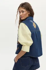 Chaleco de denim azul oscuro con cuello clásico y bordados florales en tonos amarillos, blancos y celestes en la parte delantera.
