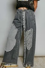 Pantalón de jean gris oscuro con corte holgado, parches asimétricos estilo patchwork con estampados de rayas y animal print en las rodillas, y detalle de tira central con botones a presión y encaje.