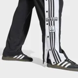 Pantalon deportivo Adidas modelo Adibreak, de corte recto y color negro. Presenta las icónicas tres tiras blancas laterales y botones a presión a lo largo de las piernas, con logo de la marca bordado en la parte inferior.