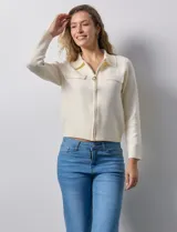 Cardigan de tejido punto color crema, con cuello tipo solapa, cierre frontal completo con tirador circular metálico y dos bolsillos de parche simulados en el frente. Presenta detalles de canalé en los hombros.