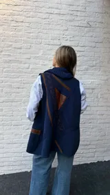 Chaleco de punto color azul marino con cuello alto tipo polera. Presenta un diseño asimétrico con detalles en cuero marrón y una franja de tejido trenzado en la espalda. Incluye bolsillos laterales con detalles en cuero.