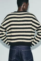 Chaqueta de punto con cuello redondo y manga larga, que combina un diseño de rayas horizontales en blanco y negro con paneles frontales decorados con motivos de crochet en tonos tierra y granate. Presenta cierre frontal con botones.