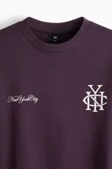 Remera de algodón color bordó con cuello redondo acanalado, mangas cortas y hombros caídos. Presenta un estampado frontal con el texto 'New York City' y un monograma entrelazado en color blanco.