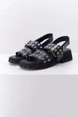 Sandalia de cuero color marrón con suela track negra. Presenta dos tiras frontales con ojalillos metálicos y una tira trasera con cierre de velcro.