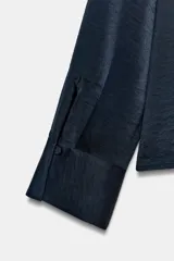Camisa de cuello solapa y manga larga, confeccionada en tejido satinado azul marino. Presenta cierre frontal con botones forrados a tono y puños rectos.