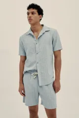 Camisa celeste a rayas verticales, de manga corta, con cuello tipo camp-collar, botones de madera y parche con logo.