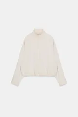 Cazadora estilo bomber de color beige, con cuello subido, manga larga y cierre frontal con cremallera. Presenta bolsillos delanteros de vivo y bajo ajustable con cordones elásticos laterales.