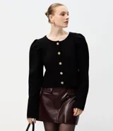 Cardigan de punto negro con cuello redondo, mangas largas abullonadas y cierre frontal con botones decorativos en forma de flor dorada. Presenta un corte corto y ajustado.