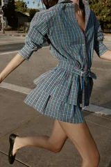 Vestido corto de manga larga con cuello solapa y cierre frontal de botones a presión. Presenta un diseño a cuadros en tonos azul y marrón, cintura ajustable con cinturón de hebilla y falda con volantes en el bajo.