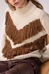 Sweater de punto color crema con cuello medio alto y silueta relajada. Presenta un diseño frontal con líneas en V decoradas con flecos largos de color marrón aplicados a mano.