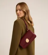 Cartera tipo shoulder bag de tamaño mediano, confeccionada en poliuretano color bordó. Presenta una solapa frontal con un detalle metálico dorado de forma orgánica y una correa ajustable con detalles decorativos.