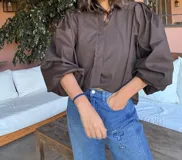 Camisa de color marrón oscuro con cuello clásico y mangas abullonadas con puños elásticos. Presenta un corte holgado y cierre frontal con botones.