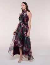 Vestido largo sin mangas de la marca RM Richards, con cuello alto halter y diseño asimétrico. Presenta un estampado floral en tonos bordó, verde y rosado sobre base satinada, con lazo ajustable en la cintura y falda fluida con capas asimétricas.
