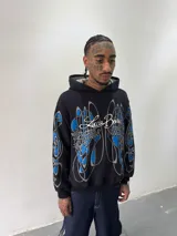 Hoodie oversize de color negro con estampado artístico en pecho y mangas en tonos azul y gris. Presenta capucha forrada con tejido a cuadros, costuras reforzadas y grifa de la marca en la manga.