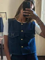 Chaleco de jean azul oscuro con botones dorados, dos bolsillos frontales con solapa y un bordado de letras en el bolsillo izquierdo.
