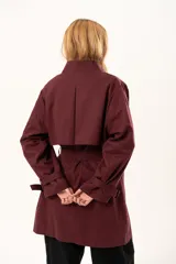 Trench de corte holgado color bordó, con cuello camisero, cierre frontal con botones y cinturón ajustable con hebilla. Presenta mangas largas con detalles de correas en los puños y bolsillos laterales.