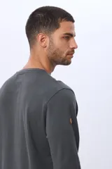 Remera de algodón texturada color gris oscuro, de manga larga, con cuello redondo y botones en la parte frontal. Presenta un pequeño logo bordado en el pecho y un parche decorativo en la manga izquierda.