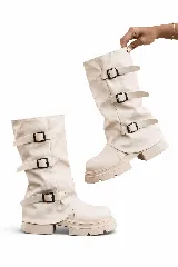 Bota biker de caña alta color crema, con suela track gruesa y tres correas ajustables con hebillas cuadradas negras en la caña. Presenta un diseño con polaina superpuesta.
