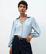 Camisa de denim liviano confeccionada en lyocell, con cuello camisero, cierre frontal de botones y mangas largas abullonadas con puños elásticos ajustados.