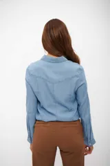 Camisa de jean en tono celeste claro, con cuello clásico, cierre frontal de botones y costuras frontales verticales que definen la silueta.