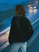 Campera de textura afelpada en color negro, con cuello redondo, cierre frontal con botones y bolsillos delanteros. Presenta detalles de terminaciones en material sintético liso a lo largo de la abertura y los bolsillos.