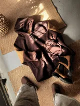 Campera con capucha de diseño patchwork, confeccionada en tejido tipo corderito en tonos marrón y tostado. Presenta un diseño que simula un paisaje montañoso con cortes irregulares y etiqueta de marca en la parte posterior.
