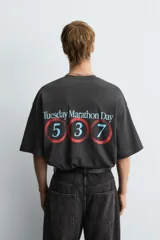 Camiseta negra con efecto lavado, corte boxy fit, cuello redondo y manga corta. Presenta un estampado frontal con texto en tonos turquesa, rojo y blanco que dice "Tuesday 600 Marathon Day Hyde Park".
