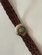 Cinto trenzado de cuero color chocolate con hebilla circular metálica grabada y piedra central.