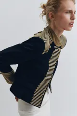 Chaqueta de estilo militar en color negro, con cuello subido y puños en terciopelo color verde oliva. Presenta detalles de alamares dorados en el frente y hombros, bolsillos con solapa y cierre frontal con botones metálicos en relieve.