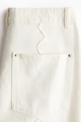 Jean de tiro regular y corte acampanado, confeccionado en denim de algodón color blanco. Presenta diseño de cinco bolsillos y detalles de costuras con ribetes bordados ondulados en el frente y los laterales.