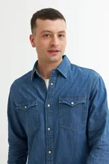 Camisa de denim azul con cuello clásico, cierre frontal con botones a presión, dos bolsillos con solapa abotonada en el pecho y mangas largas con puños abotonados.