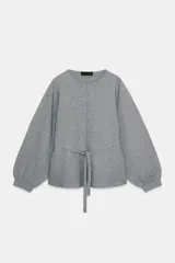 Jersey de punto color gris, con cuello redondo, mangas largas abullonadas con puños elásticos y cintura ajustable mediante cordón. Presenta una costura vertical marcada en la parte frontal.