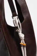 Bolso de mano y hombro grande de cuero marrón oscuro, tipo hobo, con diseño estructurado mediante costuras frontales y asa integrada. Incluye detalle lateral de cordones blancos con cuentas decorativas y el logotipo plateado de la marca en la parte inferior frontal.