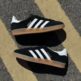 Championes Adidas Gazelle Indoor de diseño clásico, confeccionados en gamuza negra con las icónicas tres tiras laterales en blanco. Presentan el nombre del modelo en letras doradas, talón en contraste blanco y suela de goma color caramelo.