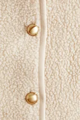 Chaqueta de sherpa suave con cuello redondo, corte holgado y cierre frontal mediante botones metálicos dorados. Presenta ribetes de cinta fina en los bordes y mangas largas.
