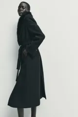 Abrigo largo de lana en color negro, con cuello de solapa desmontable en piel sintética, mangas largas con hombreras y cierre frontal con botón y cinturón ajustable del mismo tejido.