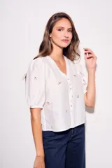Blusa de viscosa color marrón, con escote en V, mangas cortas abullonadas y cierre frontal con botones. Cuenta con pequeños detalles bordados distribuidos por la prenda.