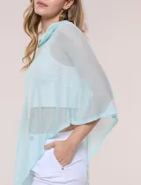 Poncho liviano de tejido de punto fino calado, color blanco, con silueta asimétrica envolvente, escote bote drapeado y mangas integradas estilo capa.