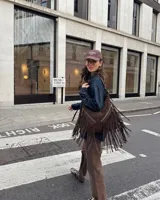 Bolso tipo hobo de cuero gamuzado color marrón oscuro, con diseño de flecos largos en el frente y dos bolsillos diagonales con cierre metálico. Cuenta con manija regulable y cierre principal metálico.