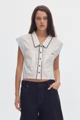 Camisa cropped sin mangas de algodón, con diseño colorblock en tonos blanco y celeste, cuello camisero con ribete negro, cierre frontal de botones y bolsillos en el pecho.