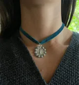 Gargantilla de cinta de terciopelo color verde azulado con dije circular plateado que presenta la imagen de la Virgen de Guadalupe rodeada de un relieve de rosas.