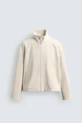 Cazadora de tejido color crema, con cuello alto, cierre frontal de cremallera y bolsillos de vivo en la cadera. Presenta un calce relajado (relaxed fit).